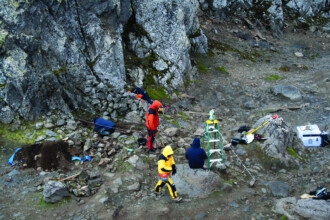 A imagem mostra uma paisagem rochosa com quatro pessoas em atividades de escavação ou pesquisa. O chão é composto de terra e pedras, e um grande paredão rochoso de cor cinza, com musgo em algumas partes, domina a parte superior da imagem. As pessoas, vestindo roupas de cores vivas como vermelho, azul e amarelo, estão dispersas pelo terreno rochoso. No centro, uma pessoa com casaco laranja está de pé. Outra, vestindo um casaco amarelo, está agachada, de costas para a câmera. Uma terceira pessoa, com agasalho azul, está agachada perto de uma fenda no solo. Vários objetos e equipamentos, incluindo uma escada verde, baldes e caixas de plástico, estão espalhados no chão, conectados por cordas brancas esticadas e presas em pedras. O nome "Renato Rabelo" está visível no canto inferior direito.