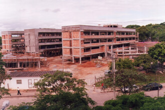 A imagem mostra uma vista externa de um canteiro de obras de um edifício de múltiplos andares. A construção principal, ao centro, é um prédio retangular de quatro andares em processo de finalização. Sua estrutura de concreto está visível, e as paredes de tijolos aparentes estão sendo erguidas. Ao fundo, à esquerda, há uma segunda estrutura de concreto, de forma similar e em estágio inicial de construção. A área ao redor dos edifícios está repleta de andaimes de madeira e materiais de construção espalhados pelo chão de terra batida. Em primeiro plano, à esquerda, há um pequeno prédio branco de apenas um andar, com um telhado baixo. Um homem, de camisa clara e calça escura, caminha em direção a ele. À direita do prédio pequeno, um portão metálico branco está parcialmente aberto. A rua em frente é de terra e há árvores e vegetação ao longo da via. Um carro escuro, possivelmente um sedã, está estacionado debaixo de uma árvore no canto inferior direito. Duas pessoas são vistas em pé na calçada, perto da entrada principal. O céu está nublado, com nuvens acinzentadas, sugerindo um tempo encoberto.