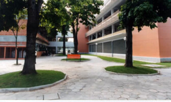 Imagem mostra um pátio externo entre edifícios, pavimentado com blocos de concreto hexagonais. O ambiente é cercado por árvores de troncos grossos e folhas verdes plantadas em canteiros elevados e arredondados. No centro do pátio, há um banco laranja de metal. Os edifícios ao redor são de múltiplos andares. À esquerda, a parede de um prédio é de tijolos à vista e possui janelas grandes. À direita, a parede de outro prédio é bege, com faixas horizontais de concreto e janelas de metal. No centro, ao fundo, os edifícios são interligados por uma passarela de concreto, e é possível ver pessoas sentadas na parte de baixo, em um espaço sombreado. A iluminação é natural, proveniente da luz do dia.
