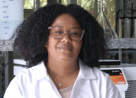 Pesquisadora Rayane Silva, mulher negra com cabelos cacheados na altura dos ombros e usa jaleco branco