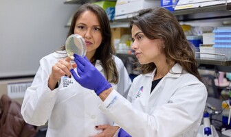 Camila Coelho e Rainna Fantin são duas mulheres vestidas de jaleco branco; ambas seguram um recipiente de vidro ou de plástico em um laboratório