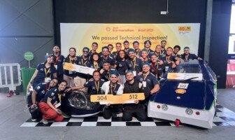 Na frente do banner amarelo da competição estão 28 alunos, todos com uniforme azul marinho e medalhas penduradas no pescoço. Eles estão ao lado de dois prótotipos de carros