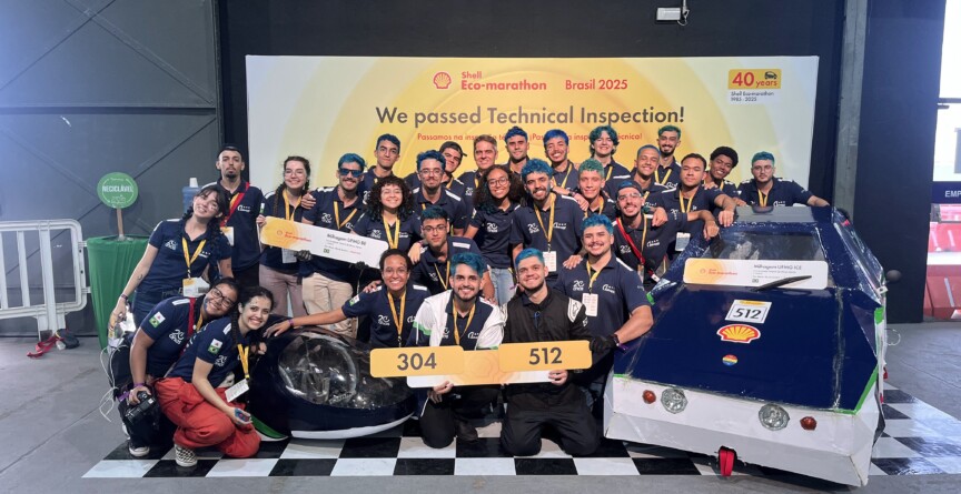Na frente do banner amarelo da competição estão 28 alunos, todos com uniforme azul marinho e medalhas penduradas no pescoço. Eles estão ao lado de dois prótotipos de carros