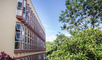 Fachada lateral da Escola de Música, cor laranja claro, com várias janelas com tom de vermelho escuro; ao lado, há árvores