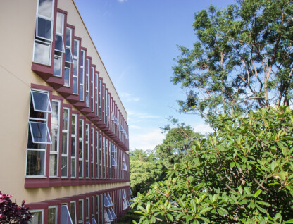 Fachada lateral da Escola de Música, cor laranja claro, com várias janelas com tom de vermelho escuro; ao lado, há árvores