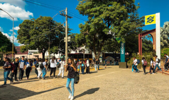 Estudantes andando em frente à Praça de Serviços; à direita, placas com logo de bancos; ao fundo, árvores e dois ônibus