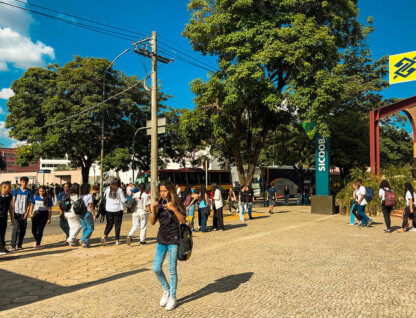 Estudantes andando em frente à Praça de Serviços; à direita, placas com logo de bancos; ao fundo, árvores e dois ônibus