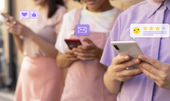 Três jovens, vestidos em tons de rosa e roxo, com celulares nas mãos. Sobre os aparelhos estão ícones de mensagem, coração, emoji e estrelas