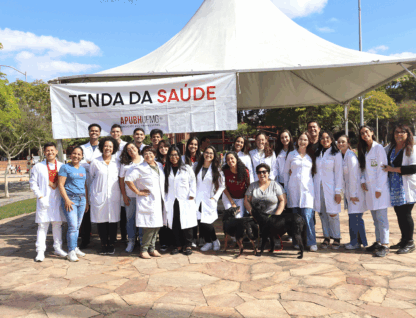 Grupo de professores e estudantes, em sua maioria vestidos com jalecos brancos, em frente a uma faixa onde se lê 'Tenda da Saúde'