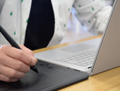 Diante de um notebook, pessoa com blusa branca faz anotações em um tablet