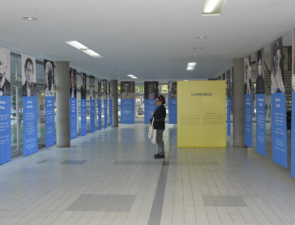 Homem de óculos, roupa preta e tiara vermelha presa ao cabelo observa retratos das personalidades homenageadas na exposição 'UFMG centenária: memoráveis'