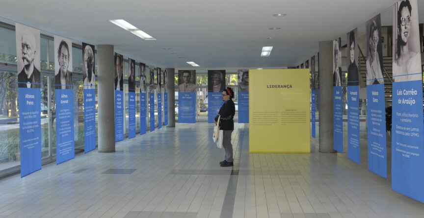 Homem de óculos, roupa preta e tiara vermelha presa ao cabelo observa retratos das personalidades homenageadas na exposição 'UFMG centenária: memoráveis'
