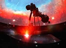 A imagem mostra o interior escuro de um planetário, com uma vasta projeção de estrelas e nebulosas coloridas em uma cúpula. Um grande projetor de estrelas, de cor escura, é visível em silhueta, posicionado no centro da imagem. A cúpula exibe tons vibrantes de vermelho e laranja na parte inferior, misturando-se com azuis e brancos na parte superior, simulando o espaço sideral. Em primeiro plano, uma plataforma metálica com orifícios e uma luz vermelha brilhante irradiando para cima é visível, possivelmente um laser ou outro efeito de projeção.