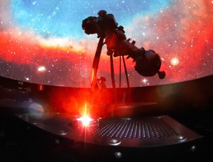 A imagem mostra o interior escuro de um planetário, com uma vasta projeção de estrelas e nebulosas coloridas em uma cúpula. Um grande projetor de estrelas, de cor escura, é visível em silhueta, posicionado no centro da imagem. A cúpula exibe tons vibrantes de vermelho e laranja na parte inferior, misturando-se com azuis e brancos na parte superior, simulando o espaço sideral. Em primeiro plano, uma plataforma metálica com orifícios e uma luz vermelha brilhante irradiando para cima é visível, possivelmente um laser ou outro efeito de projeção.