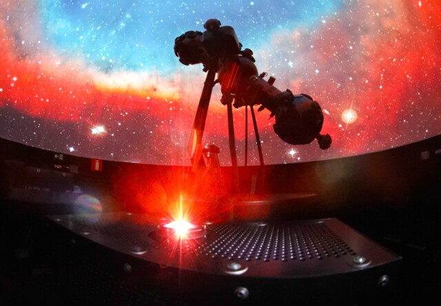 A imagem mostra o interior escuro de um planetário, com uma vasta projeção de estrelas e nebulosas coloridas em uma cúpula. Um grande projetor de estrelas, de cor escura, é visível em silhueta, posicionado no centro da imagem. A cúpula exibe tons vibrantes de vermelho e laranja na parte inferior, misturando-se com azuis e brancos na parte superior, simulando o espaço sideral. Em primeiro plano, uma plataforma metálica com orifícios e uma luz vermelha brilhante irradiando para cima é visível, possivelmente um laser ou outro efeito de projeção.