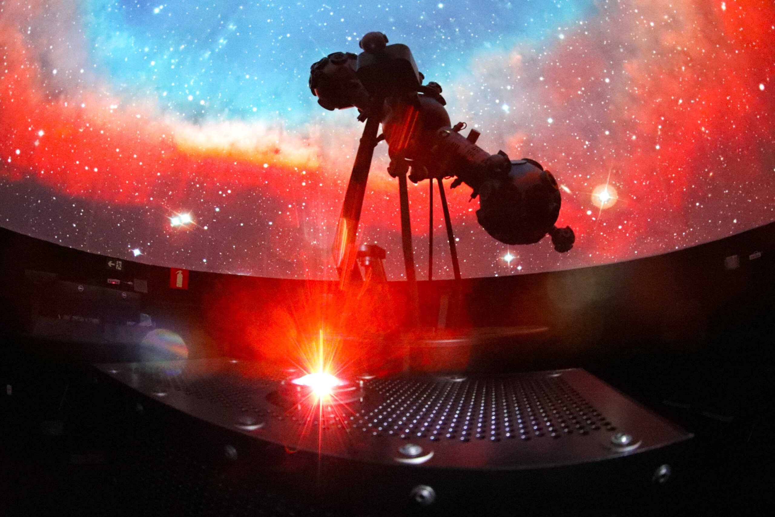 A imagem mostra o interior escuro de um planetário, com uma vasta projeção de estrelas e nebulosas coloridas em uma cúpula. Um grande projetor de estrelas, de cor escura, é visível em silhueta, posicionado no centro da imagem. A cúpula exibe tons vibrantes de vermelho e laranja na parte inferior, misturando-se com azuis e brancos na parte superior, simulando o espaço sideral. Em primeiro plano, uma plataforma metálica com orifícios e uma luz vermelha brilhante irradiando para cima é visível, possivelmente um laser ou outro efeito de projeção.