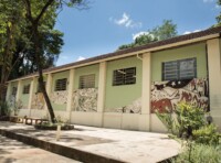 Imagem mostra edifício de um andar com fachada pintada em tons de verde claro e branco, sob um céu azul claro com algumas nuvens. O telhado é formado por telhas de cerâmica escuras. A parede externa do edifício é decorada com grandes murais em tons de verde, branco e vermelho escuro, retratando figuras estilizadas e abstratas. Várias janelas retangulares com grades são visíveis na fachada. Em frente ao prédio, há um pátio de concreto com algumas plantas e um banco longo de cor clara. À esquerda do edifício, uma árvore grande com tronco e galhos ramificados se eleva, e sua folhagem verde escura se destaca contra o céu