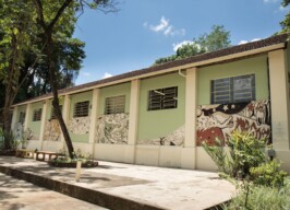 Imagem mostra edifício de um andar com fachada pintada em tons de verde claro e branco, sob um céu azul claro com algumas nuvens. O telhado é formado por telhas de cerâmica escuras. A parede externa do edifício é decorada com grandes murais em tons de verde, branco e vermelho escuro, retratando figuras estilizadas e abstratas. Várias janelas retangulares com grades são visíveis na fachada. Em frente ao prédio, há um pátio de concreto com algumas plantas e um banco longo de cor clara. À esquerda do edifício, uma árvore grande com tronco e galhos ramificados se eleva, e sua folhagem verde escura se destaca contra o céu