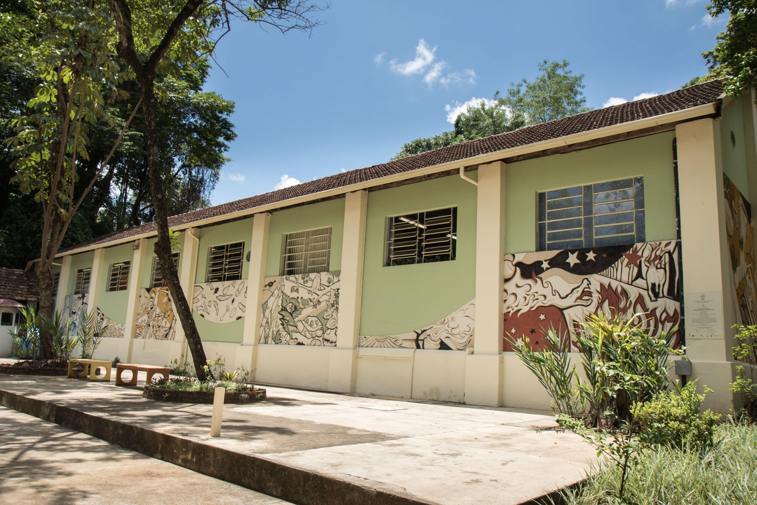 Imagem mostra edifício de um andar com fachada pintada em tons de verde claro e branco, sob um céu azul claro com algumas nuvens. O telhado é formado por telhas de cerâmica escuras. A parede externa do edifício é decorada com grandes murais em tons de verde, branco e vermelho escuro, retratando figuras estilizadas e abstratas. Várias janelas retangulares com grades são visíveis na fachada. Em frente ao prédio, há um pátio de concreto com algumas plantas e um banco longo de cor clara. À esquerda do edifício, uma árvore grande com tronco e galhos ramificados se eleva, e sua folhagem verde escura se destaca contra o céu