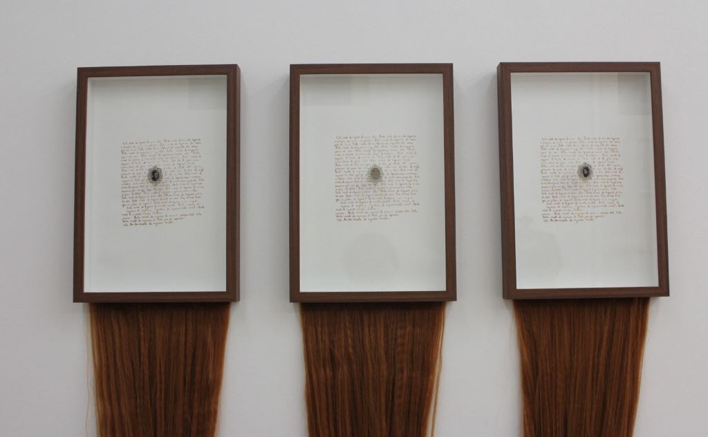 A imagem mostra três obras de arte semelhantes, dispostas lado a lado em uma parede branca. Cada obra consiste em um quadro retangular com moldura marrom, contendo um papel com texto manuscrito em tinta escura, e um medalhão ou objeto circular escuro no centro. Abaixo de cada quadro, uma longa cortina de cabelo humano castanho-avermelhado desce verticalmente, pendurada na parte inferior da moldura. As mechas de cabelo são lisas e brilhantes, formando um contraste textural com a superfície branca da parede.