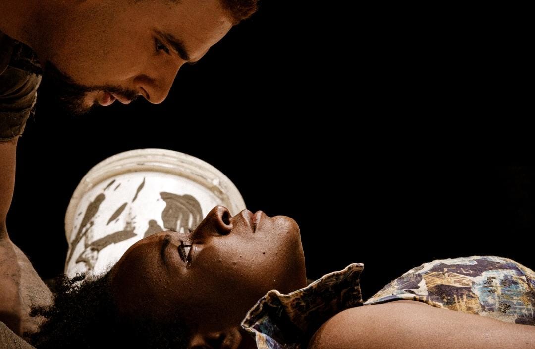 A imagem mostra um close-up de duas pessoas em uma interação. Uma pessoa está deitada, com a cabeça levemente erguida e o rosto escuro, olhando para cima. Ela veste uma blusa estampada em tons terrosos. Acima dela, outra pessoa, com pele mais clara e barba, está inclinada sobre a primeira, olhando para baixo. Há um balde branco com resíduos escuros sobre a cabeça da pessoa deitada. O fundo da imagem é totalmente preto, criando um contraste dramático e focando a atenção nas duas figuras.