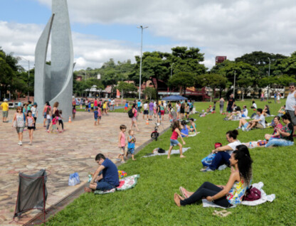 A imagem mostra um parque ou praça com uma grande escultura geométrica de cor clara à esquerda. O local está cheio de pessoas, incluindo adultos e crianças, desfrutando de um dia ao ar livre. Uma vasta área gramada se estende pelo lado direito, onde muitas pessoas estão sentadas ou deitadas, relaxando. Há também uma área pavimentada com pedras no lado esquerdo, onde mais pessoas estão circulando. O fundo é preenchido por árvores e edifícios, sob um céu parcialmente nublado.