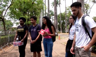 A imagem mostra um grupo de cinco jovens, três mulheres e dois homens, em pé em um ambiente ao ar livre, possivelmente um campus universitário ou parque, com muitas árvores ao fundo. Eles estão de frente para a câmera, alguns sorrindo. A jovem no centro, com cabelo encaracolado e comprido, veste uma blusa rosa e calça jeans, e segura um objeto vermelho. À esquerda, uma mulher de pele escura veste uma camiseta preta e segura um pacote de bebidas. Os outros jovens vestem camisetas casuais e alguns usam mochilas. O chão é de concreto e há grades metálicas no lado esquerdo.