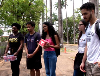 A imagem mostra um grupo de cinco jovens, três mulheres e dois homens, em pé em um ambiente ao ar livre, possivelmente um campus universitário ou parque, com muitas árvores ao fundo. Eles estão de frente para a câmera, alguns sorrindo. A jovem no centro, com cabelo encaracolado e comprido, veste uma blusa rosa e calça jeans, e segura um objeto vermelho. À esquerda, uma mulher de pele escura veste uma camiseta preta e segura um pacote de bebidas. Os outros jovens vestem camisetas casuais e alguns usam mochilas. O chão é de concreto e há grades metálicas no lado esquerdo.