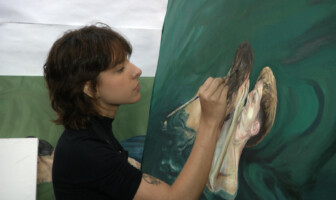 A imagem mostra uma mulher jovem com cabelo castanho-avermelhado e curto, concentrada em uma pintura a óleo em uma tela grande. Ela está de perfil, usando uma blusa preta de manga comprida e um colar preto. A mulher segura um pincel na mão direita, aplicando tinta em uma parte da pintura que retrata um rosto com cabelo fluindo. A pintura em tons de verde escuro e creme parece representar figuras submersas ou em movimento. O fundo da imagem é um estúdio de arte, com uma parede clara e outras obras de arte parcialmente visíveis.