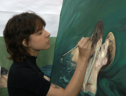 A imagem mostra uma mulher jovem com cabelo castanho-avermelhado e curto, concentrada em uma pintura a óleo em uma tela grande. Ela está de perfil, usando uma blusa preta de manga comprida e um colar preto. A mulher segura um pincel na mão direita, aplicando tinta em uma parte da pintura que retrata um rosto com cabelo fluindo. A pintura em tons de verde escuro e creme parece representar figuras submersas ou em movimento. O fundo da imagem é um estúdio de arte, com uma parede clara e outras obras de arte parcialmente visíveis.