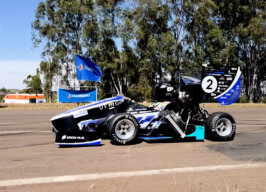 A imagem mostra um carro de corrida monoposto, de design aerodinâmico, parado em uma pista asfaltada sob um céu azul claro. O carro é predominantemente preto, com detalhes em azul-claro, branco e verde-limão, e o número "2" em branco na parte traseira. Vários logotipos de patrocinadores são visíveis na carroceria. Um piloto está sentado no cockpit, usando capacete. Ao fundo, uma fileira de árvores altas, bandeiras azuis da "SAE BRASIL" e estruturas de barraca são visíveis. Há também uma ambulância branca e laranja e alguns cones de tráfego.