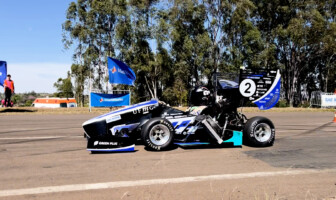 A imagem mostra um carro de corrida monoposto, de design aerodinâmico, parado em uma pista asfaltada sob um céu azul claro. O carro é predominantemente preto, com detalhes em azul-claro, branco e verde-limão, e o número "2" em branco na parte traseira. Vários logotipos de patrocinadores são visíveis na carroceria. Um piloto está sentado no cockpit, usando capacete. Ao fundo, uma fileira de árvores altas, bandeiras azuis da "SAE BRASIL" e estruturas de barraca são visíveis. Há também uma ambulância branca e laranja e alguns cones de tráfego.