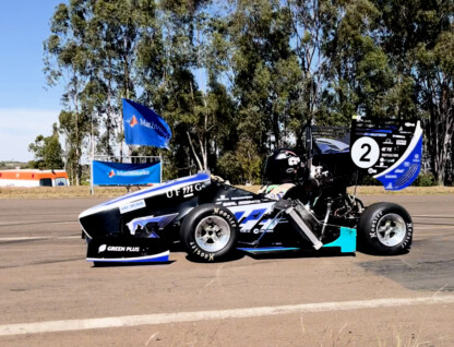 A imagem mostra um carro de corrida monoposto, de design aerodinâmico, parado em uma pista asfaltada sob um céu azul claro. O carro é predominantemente preto, com detalhes em azul-claro, branco e verde-limão, e o número "2" em branco na parte traseira. Vários logotipos de patrocinadores são visíveis na carroceria. Um piloto está sentado no cockpit, usando capacete. Ao fundo, uma fileira de árvores altas, bandeiras azuis da "SAE BRASIL" e estruturas de barraca são visíveis. Há também uma ambulância branca e laranja e alguns cones de tráfego.