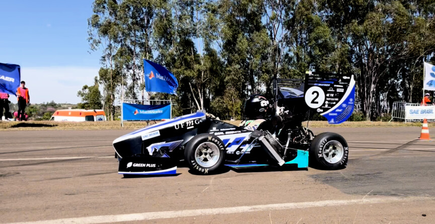 A imagem mostra um carro de corrida monoposto, de design aerodinâmico, parado em uma pista asfaltada sob um céu azul claro. O carro é predominantemente preto, com detalhes em azul-claro, branco e verde-limão, e o número "2" em branco na parte traseira. Vários logotipos de patrocinadores são visíveis na carroceria. Um piloto está sentado no cockpit, usando capacete. Ao fundo, uma fileira de árvores altas, bandeiras azuis da "SAE BRASIL" e estruturas de barraca são visíveis. Há também uma ambulância branca e laranja e alguns cones de tráfego.