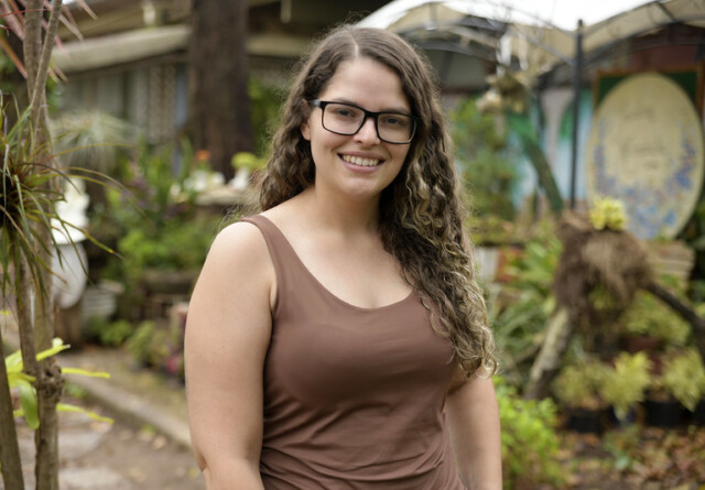 Caroline Viana é uma mulher jovem de cabelos compridos castanhos. Ela usa um par de óculos marrom. Ela está em uma área de jardins na Faculdade de Educação