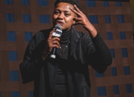 A imagem mostra uma pessoa de pele escura com cabelo curto e crespo, em pé, segurando um microfone e gesticulando com a mão esquerda, que faz um sinal de "V" com os dedos. A pessoa está sorrindo e olhando para o lado direito da foto. Ela veste uma jaqueta ou casaco preto sobre uma blusa de gola alta preta, e calças claras com padrões. Um colar com um pingente em forma de cruz é visível. O fundo da imagem é uma parede com um padrão de quadrados marrons e azuis, e uma faixa vermelha na parte superior.