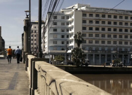 A imagem mostra uma paisagem urbana com um rio ou canal em primeiro plano à direita, e uma ponte de concreto com um parapeito robusto se estendendo pelo lado esquerdo. Pessoas caminham sobre a ponte. Ao fundo, uma fileira de edifícios de arquitetura variada domina a paisagem. Um grande edifício branco com muitas janelas se destaca no centro, e à sua frente, algumas palmeiras são visíveis. Mais ao fundo e à direita, outros edifícios altos completam o cenário da cidade. Fios elétricos se cruzam no céu, e um ônibus azul e amarelo é visível na rua à esquerda.