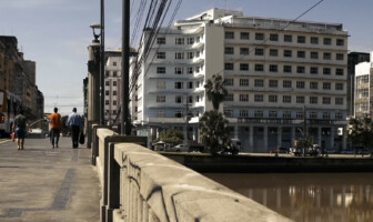 A imagem mostra uma paisagem urbana com um rio ou canal em primeiro plano à direita, e uma ponte de concreto com um parapeito robusto se estendendo pelo lado esquerdo. Pessoas caminham sobre a ponte. Ao fundo, uma fileira de edifícios de arquitetura variada domina a paisagem. Um grande edifício branco com muitas janelas se destaca no centro, e à sua frente, algumas palmeiras são visíveis. Mais ao fundo e à direita, outros edifícios altos completam o cenário da cidade. Fios elétricos se cruzam no céu, e um ônibus azul e amarelo é visível na rua à esquerda.