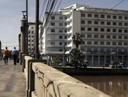 A imagem mostra uma paisagem urbana com um rio ou canal em primeiro plano à direita, e uma ponte de concreto com um parapeito robusto se estendendo pelo lado esquerdo. Pessoas caminham sobre a ponte. Ao fundo, uma fileira de edifícios de arquitetura variada domina a paisagem. Um grande edifício branco com muitas janelas se destaca no centro, e à sua frente, algumas palmeiras são visíveis. Mais ao fundo e à direita, outros edifícios altos completam o cenário da cidade. Fios elétricos se cruzam no céu, e um ônibus azul e amarelo é visível na rua à esquerda.