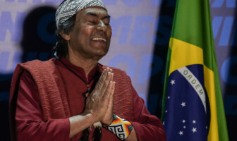 Ailton Krenak, ativista do movimento socioambiental e defensor dos direitos dos povos indígenas, está à esquerda da foto sorrindo. Tem os olhos fechados e as mãos juntas, como se em oração. Na direita da foto, um pouco mais ao fundo, uma bandeira do Brasil.
