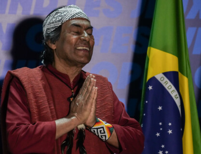 Ailton Krenak, ativista do movimento socioambiental e defensor dos direitos dos povos indígenas, está à esquerda da foto sorrindo. Tem os olhos fechados e as mãos juntas, como se em oração. Na direita da foto, um pouco mais ao fundo, uma bandeira do Brasil.