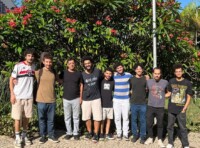 A imagem mostra um grupo de oito homens, em pé lado a lado, sorrindo e posando para a câmera. Eles estão em um ambiente ao ar livre, com um grande arbusto florido em vermelho atrás deles. O grupo exibe uma variedade de tons de pele e estilos de cabelo. Eles vestem roupas casuais, incluindo camisetas, calças jeans e bermudas. Um dos homens carrega um case de instrumento musical nas costas. O chão é pavimentado com pedras claras e o sol ilumina a cena.