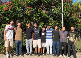 A imagem mostra um grupo de oito homens, em pé lado a lado, sorrindo e posando para a câmera. Eles estão em um ambiente ao ar livre, com um grande arbusto florido em vermelho atrás deles. O grupo exibe uma variedade de tons de pele e estilos de cabelo. Eles vestem roupas casuais, incluindo camisetas, calças jeans e bermudas. Um dos homens carrega um case de instrumento musical nas costas. O chão é pavimentado com pedras claras e o sol ilumina a cena.