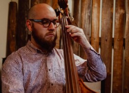 A imagem mostra um homem careca com barba e óculos de armação escura, concentrado enquanto toca um contrabaixo acústico. Ele veste uma camisa de manga comprida com um padrão paisley em tons de rosa e roxo. O homem está com os olhos fechados ou semicerrados, e suas mãos estão posicionadas nas cordas do instrumento de madeira. O fundo da imagem é uma parede de madeira com tábuas verticais, com iluminação suave, criando um ambiente de estúdio ou palco.