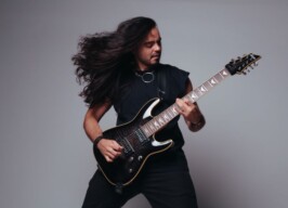 A imagem mostra um jovem com cabelos longos e escuros, que estão voando para a esquerda, simulando movimento. Ele está vestido com uma camiseta preta sem mangas, calças escuras e usa um colar prateado. O jovem está tocando uma guitarra elétrica escura com as duas mãos, e parece estar no meio de uma performance. O fundo da imagem é um cinza claro e uniforme, destacando a figura do músico.