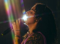 A imagem mostra uma pessoa de perfil, com cabelos longos e cacheados, cantando em um microfone. A luz forte incide diretamente sobre o rosto e o microfone, criando um brilho intenso e raios de luz coloridos, semelhantes a um arco-íris, que atravessam a imagem. A pessoa tem várias tatuagens visíveis no braço e ombro, incluindo uma rosa grande e outras menores. Ela veste uma blusa ou top de cor roxa. O fundo da imagem é escuro e desfocado, direcionando o foco para a pessoa e a luz.