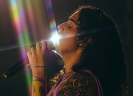A imagem mostra uma pessoa de perfil, com cabelos longos e cacheados, cantando em um microfone. A luz forte incide diretamente sobre o rosto e o microfone, criando um brilho intenso e raios de luz coloridos, semelhantes a um arco-íris, que atravessam a imagem. A pessoa tem várias tatuagens visíveis no braço e ombro, incluindo uma rosa grande e outras menores. Ela veste uma blusa ou top de cor roxa. O fundo da imagem é escuro e desfocado, direcionando o foco para a pessoa e a luz.