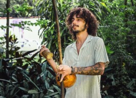 A imagem mostra um jovem com cabelos cacheados e barba, usando uma camisa listrada clara de manga curta, tocando um berimbau. Ele tem tatuagens nos braços e brincos nas orelhas. O jovem segura o berimbau na vertical, com as mãos posicionadas para produzir som. O instrumento tem uma cabaça clara na parte inferior e uma vara de madeira. O fundo da imagem é preenchido por densa vegetação verde, com folhas grandes e pequenas, sugerindo um ambiente ao ar livre.