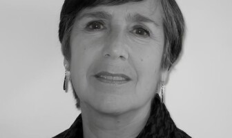 A imagem é um retrato em preto e branco de uma mulher de meia-idade ou mais velha, vista do peito para cima, olhando ligeiramente para a direita da câmera. Ela tem cabelo escuro e curto, com franja. A mulher usa brincos compridos e um lenço ou echarpe com padrão xadrez em tons escuros. Ela tem um sorriso sutil e o fundo da imagem é uma parede clara e uniforme.