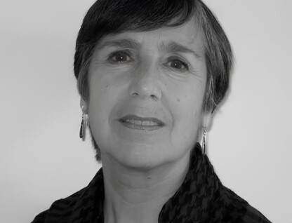 A imagem é um retrato em preto e branco de uma mulher de meia-idade ou mais velha, vista do peito para cima, olhando ligeiramente para a direita da câmera. Ela tem cabelo escuro e curto, com franja. A mulher usa brincos compridos e um lenço ou echarpe com padrão xadrez em tons escuros. Ela tem um sorriso sutil e o fundo da imagem é uma parede clara e uniforme.